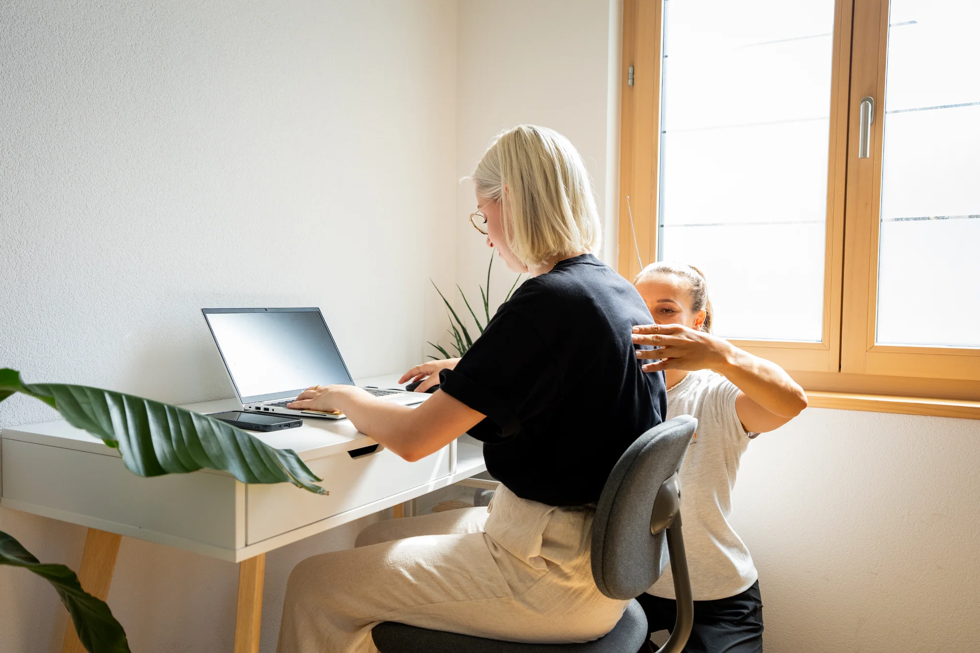 Physiotherapeutin berät einen Büroangestellten an seinem Schreibtisch; höhenverstellbarer Tisch, ergonomischer Stuhl; heller, moderner Büroraum; freundliche, professionelle Interaktion