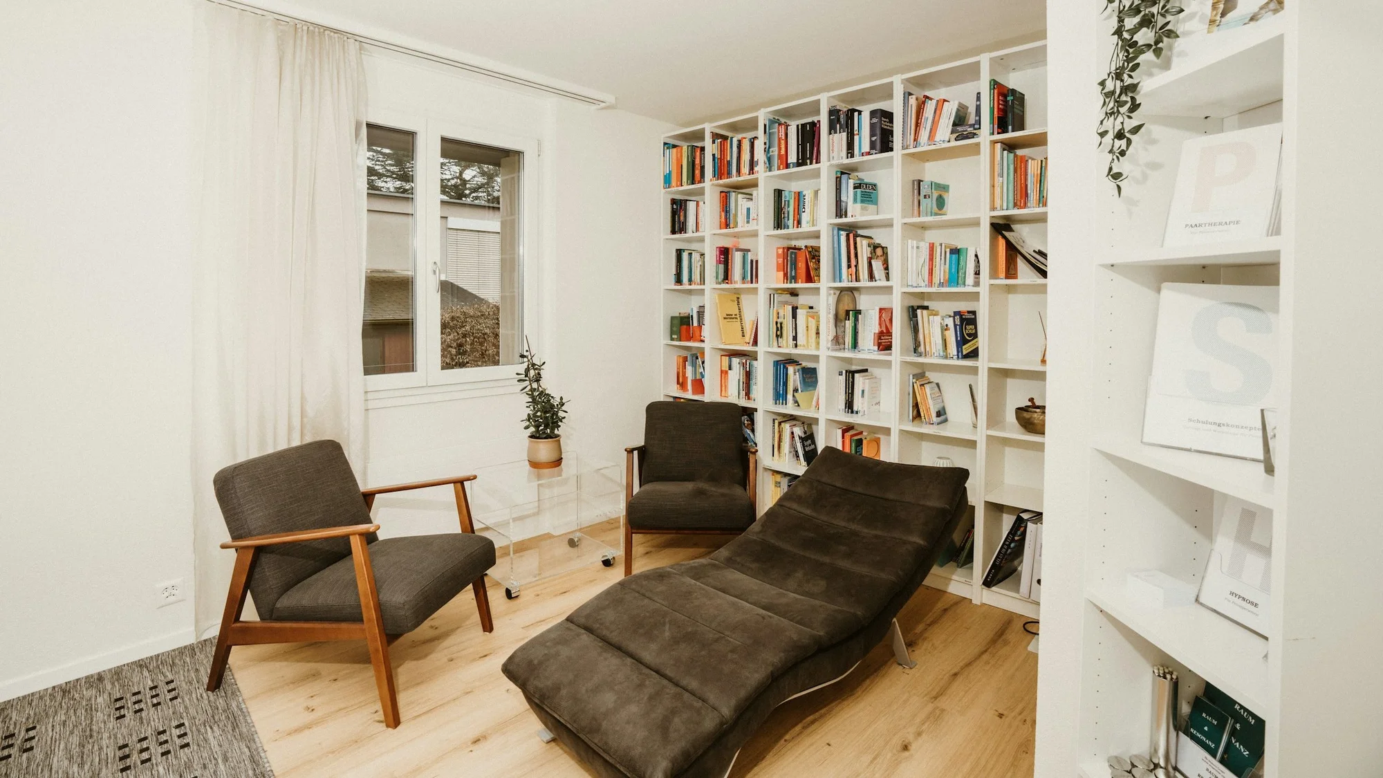 Heller Therapieraum mit zwei Sesseln, Relaxliege, Bücherwand und Pflanze am Fenster; warmes Holz, natürliches Tageslicht, keine Personen im Bild